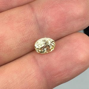 以下が含まれることがあります： 明るい黄色のオーバルカットの宝石。宝石は指の間に挟まれ、その透明度とファセットを示しています。石は暖かく金色の色合いで、光を反射します。この宝石はジュエリー作りに適しています。