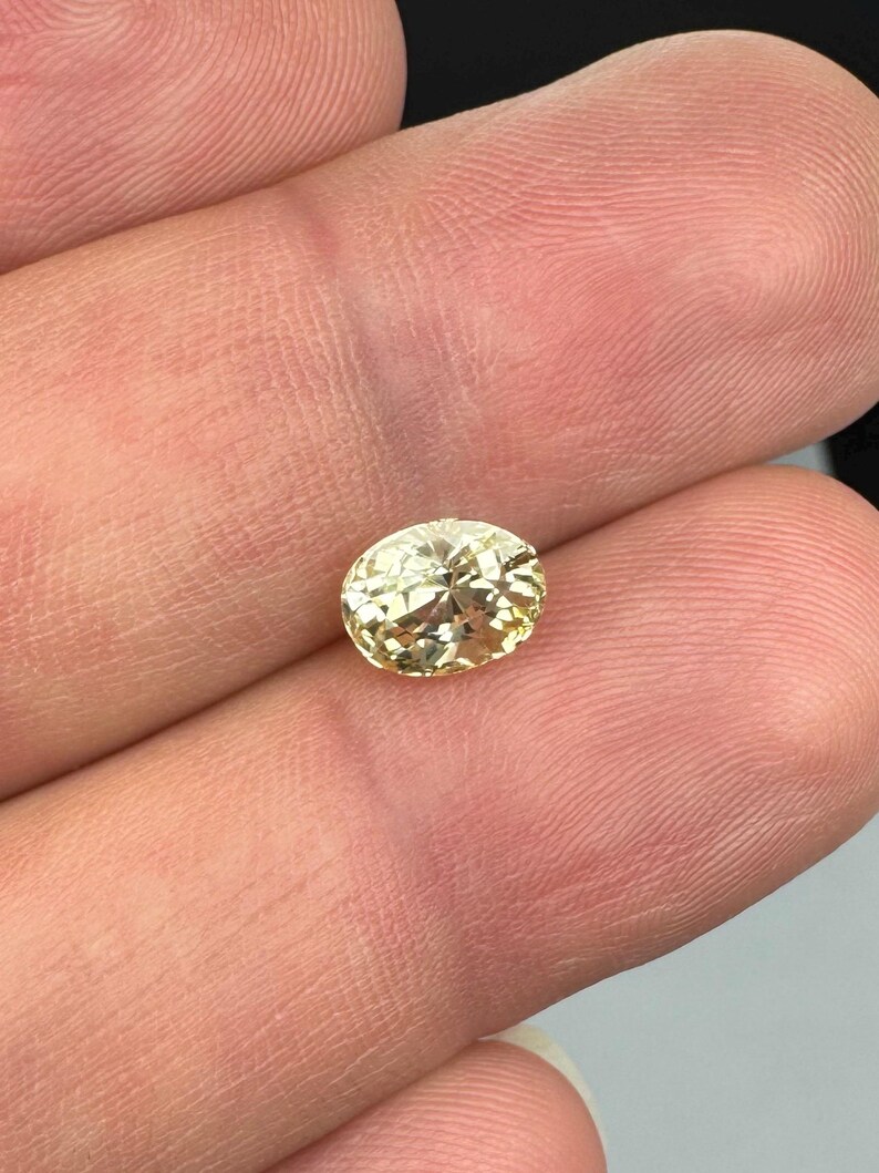 以下が含まれることがあります： 明るい黄色のオーバルカットの宝石。宝石は指の間に挟まれ、その透明度とファセットを示しています。石は暖かく金色の色合いで、光を反射します。この宝石はジュエリー作りに適しています。