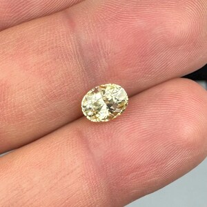 以下が含まれることがあります： 楕円形、淡い黄色の宝石。指の間に宝石が挟まれ、その形と色を示しています。石は光を反射するファセット加工された表面を持っています。背景はニュートラルな色調で、宝石の輝きを際立たせています。
