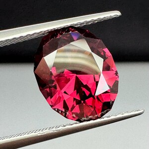 Könnte beinhalten: Ein facettierter, oval geformter Edelstein in tiefroter Farbe wird von einer silbernen Pinzette gehalten. Die vielen Facetten des Edelsteins reflektieren das Licht und erzeugen einen funkelnden Effekt. Der Hintergrund ist ein Farbverlauf von Schwarz und Grau.