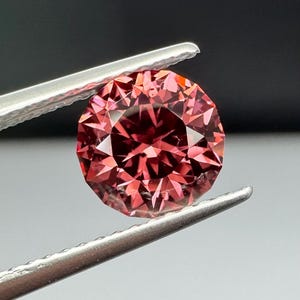 May include: A faceted, round-cut gemstone held by silver tweezers. The gem is a deep pink color with multiple facets reflecting light. The background is a gradient of black and gray.