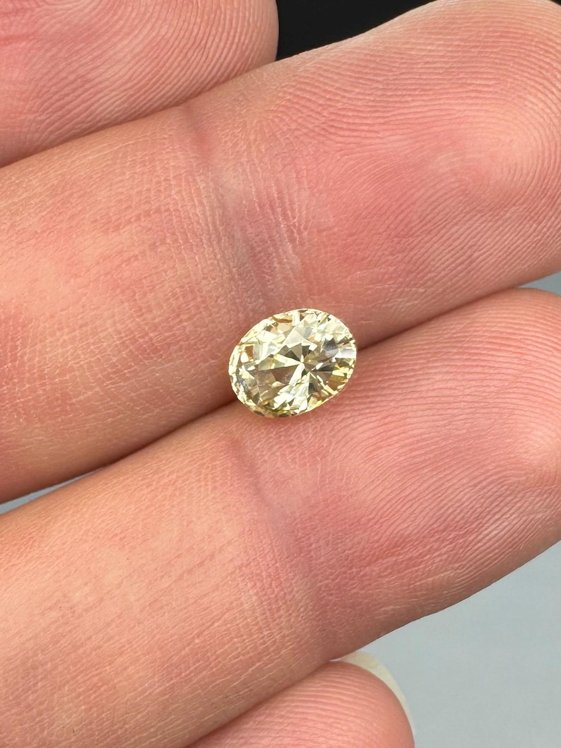 以下が含まれることがあります： 淡い黄色のオーバルカットダイヤモンド。宝石は指の間に挟まれ、そのファセットと輝きを際立たせています。ダイヤモンドの色は淡く温かみのある黄色で、カットが良く、光を反射しているようです。全体的な印象は、高品質の宝石です。
