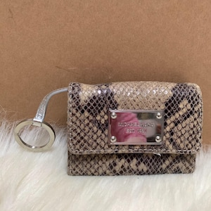 May include: A small, rectangular wallet with a snakeskin pattern in shades of brown and tan. It features a silver metal plate with the text "MICHAEL KORS EST. 1981" and a silver key ring attached.