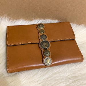 May include: A brown leather wallet with a flap closure. The wallet is adorned with a decorative strip of antique-style metal buttons and coins. The wallet is rectangular and appears to be made of genuine leather.