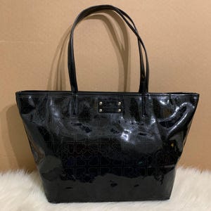 May include: Black patent leather tote bag with two top handles. The bag has a perforated design and a small gold-tone logo plate. The bag is on a white, fluffy surface.