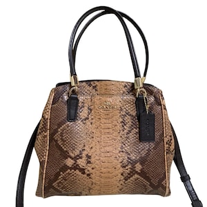 May include: A Coach handbag with a snakeskin pattern in shades of brown and tan. The bag has black handles and a black shoulder strap. Gold-colored hardware accents the bag, and a black tag hangs from the handle.