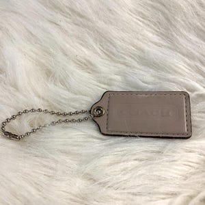 May include: A rectangular light beige leather luggage tag featuring a debossed Coach logo on the front surface. The tag is attached to a silver-toned metal ball chain, which is looped through a reinforced metal grommet on the left side of the tag.