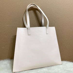 May include: A light pink tote bag with white handles. The bag has a rectangular shape with a flat bottom and a smooth surface. The handles are attached to the top of the bag with small rectangular pieces. The text "BETWEEN TWIN STARS" is printed on the front.
