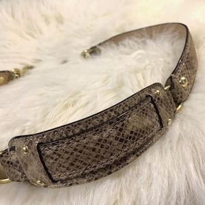 Peut inclure: Une bandoulière de sac à main incurvée présentant un motif texturé en peau de serpent dans des tons marron et beige. La sangle est agrémentée de rivets en métal doré poli et de fixations aux extrémités, avec un design structuré et des bords cousus.