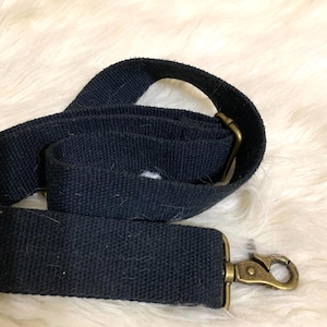 May include: A black woven fabric shoulder strap featuring an adjustable antique brass-toned metal slider and a matching swivel lobster clasp hook at the end. The strap is made of a durable, textured material and is designed for attaching to bags.
