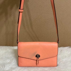 May include: A peach-colored crossbody bag with a flap closure. The bag features a black spade-shaped logo on the front and a long, adjustable strap. The bag is made of textured leather and has a simple, elegant design.