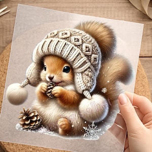 Könnte beinhalten: Eine gedruckte Illustration eines braunen Eichhörnchens mit einer beigen Strickmütze mit Ohrenklappen und Bommeln, das einen kleinen Tannenzapfen hält. Das Bild zeigt einen größeren Tannenzapfen an der Basis, weiße Schneeflocken und den Text "machs selbst" in Hellrosa.