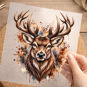 Könnte beinhalten: Ein quadratisches Transferblatt mit einer detaillierten Illustration eines Hirschkopfes mit großem Geweih. Das Kunstwerk verwendet braune, beige und dunkelblaue Spritzer und Tropfen im Aquarellstil als Hintergrundelement hinter dem Tierporträt.