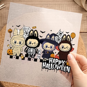 Könnte beinhalten: Ein transparenter Transferbogen mit einer Cartoon-Illustration von vier Charakteren in Halloween-Kostümen, darunter eine Mumie, ein Skelett, eine Hexe und ein Vampir, mit Spinnweben, Fledermäusen, Kürbissen und dem Text HAPPY HALLOWEEN.