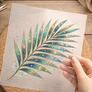 Könnte beinhalten: Ein quadratisches, halbtransparentes Transferblatt mit einer detaillierten Palmblattgrafik. Das Blattdesign ist in einem Farbverlauf aus blaugrünem, blauem und goldenem Glitzer gehalten und mit kleinen, verstreuten blauen Strasssteinen akzentuiert.