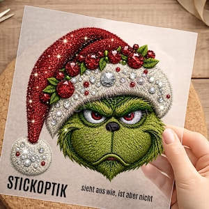Könnte beinhalten: Eine quadratische Karte mit einer gestickten grünen Kreatur mit roten Augen, die eine rot-weiße Weihnachtsmütze mit roten und klaren Kristallen trägt. Der Text STICKOPTIK und sieht aus wie, ist aber nicht ist unten in Schwarz gedruckt.