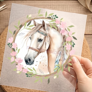 Könnte beinhalten: Aquarell-Illustration eines weißen Pferdekopfes, verziert mit einem dekorativen Zaumzeug und einem floralen Kranz. Der Kranz zeigt rosa Blüten und grüne Blätter, die ein zartes und künstlerisches Design schaffen. Mähne und Stirnlocke des Pferdes sind lang und fließend.