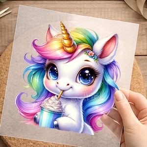 Könnte beinhalten: Eine skurrile Illustration eines Cartoon-Einhorns mit Regenbogenmähne und -schwanz, einem goldenen Horn und großen blauen Augen. Das Einhorn hält ein blau-weißes Getränk mit Schlagsahne und einem Strohhalm. Das Bild befindet sich auf einem Stück Papier.
