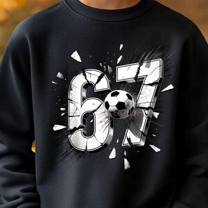 Könnte beinhalten: Schwarzes Sweatshirt mit einem weißen Grafikdesign der Zahlen "67" und einem Fußball in der Mitte. Die Zahlen scheinen auseinanderzubrechen, mit Splittern darum herum. Getragen von einer Person.