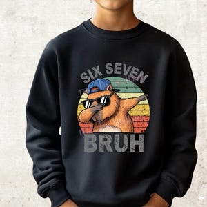 Könnte beinhalten: Schwarzes Sweatshirt mit einem Cartoon-Biber, der eine Sonnenbrille und eine blaue Kappe trägt und dabbt. Das Design enthält den Text "SIX SEVEN BRUH" in einer verwitterten Schriftart, vor einem Retro-Sonnenuntergangshintergrund.