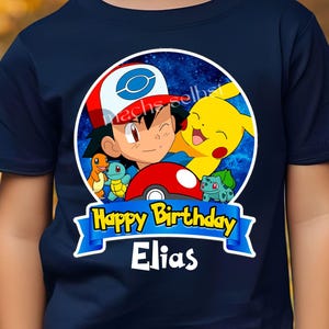 Könnte beinhalten: Ein dunkelblaues T-Shirt mit einem kreisförmigen Motiv von Ash Ketchum, Pikachu, Glumanda, Schiggy und Bisasam vor einem Sternenhintergrund. Darunter befindet sich ein blaues Banner mit dem Text Happy Birthday und dem Namen Elias.