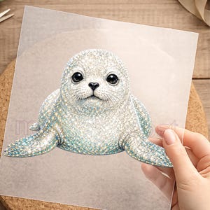 Könnte beinhalten: Detaillierte Illustration eines Seehundbabys mit glitzernder, strukturierter Optik. Der Seehund hat große, dunkle Augen und eine kleine, schwarze Nase. Sein Körper ist hauptsächlich weiß mit blauen Akzenten, auf einer transparenten Folie.