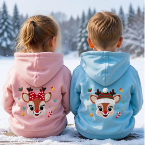 Könnte beinhalten: Ein hellrosa Kapuzenpullover und ein hellblauer Kapuzenpullover, jeweils mit einem strukturierten, gestickten Rentiergesicht auf der Rückseite. Das Rentier auf dem rosa Pullover trägt eine rote Schleife mit weißen Punkten, das auf dem blauen eine Wintermütze.