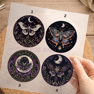 Könnte beinhalten: Vier runde Aufkleber mit Mottenillustrationen. Jeder Aufkleber hat ein anderes Motten-Design mit Details wie einem Halbmond, Sternen und floralen Elementen. Die Farbpalette umfasst Schattierungen von Lila, Blau und Weiß.