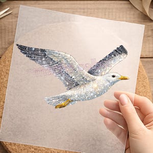 Könnte beinhalten: Ein glitzerndes, glitzerndes Möwenmotiv im Flug, mit weit ausgebreiteten Flügeln. Der Vogel ist überwiegend weiß mit grauen und schwarzen Flügelspitzen und einem gelben Schnabel und Füßen. Das Bild befindet sich auf einer transparenten Folie.