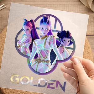 Könnte beinhalten: Ein durchsichtiger Aufkleber mit drei animierten Figuren mit violettem und blauem Haar, die futuristische Outfits in Weiß und Gold tragen. Die Figuren sind vor einem kreisförmigen violetten Emblem platziert. Das Wort GOLDEN ist unten in großen, metallisch wirkenden Buchstaben aufgedruckt.