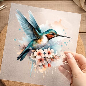 Könnte beinhalten: Ein Aquarelldruck mit einem Kolibri mit türkis, blauen und orangefarbenen Federn, die Flügel sind ausgebreitet. Der Vogel sitzt über einem Strauß rosa und weißer Blumen. Der Text "ens selbst" ist sichtbar.