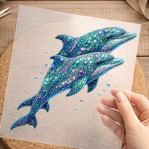 Könnte beinhalten: Detaillierte Illustration von zwei Delfinen in Blau- und Türkistönen, verziert mit juwelenartigen Verzierungen. Das Kunstwerk befindet sich auf einer transparenten Folie, mit kleinen Blasen um die Delfine.