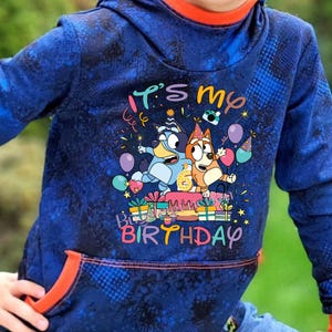 Könnte beinhalten: Blauer Kapuzenpullover mit orangefarbenem Innenfutter und Taschenbesatz. Auf der Vorderseite befindet sich eine farbenfrohe Grafik mit dem Text "It's my Birthday" sowie Zeichentrickfiguren, Luftballons und Geschenken.