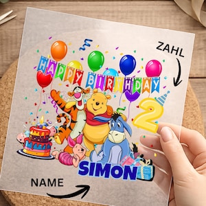 Könnte beinhalten: Eine transparente Folie mit einem farbenfrohen Geburtstagsdesign mit Winnie-the-Pooh-Figuren, Luftballons und einer Torte. Die Wörter "HAPPY BIRTHDAY" und "SIMON" sind abgebildet. Eine große gelbe Zahl "2" ist ebenfalls vorhanden.