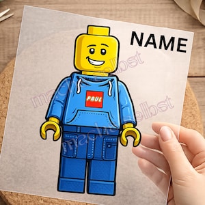 Könnte beinhalten: Ein transparenter Aufkleber mit einer Cartoon-Spielfigur mit gelbem Kopf, die einen blauen Kapuzenpullover und eine blaue Hose trägt. Auf der Brust des Pullovers befindet sich ein rotes Quadrat mit dem Namen PAUL in Weiß. Oben steht NAME in Schwarz.