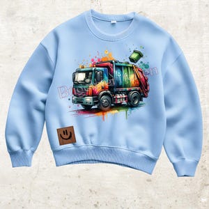 Könnte beinhalten: Hellblaues Sweatshirt mit einer farbenfrohen Illustration eines Müllwagens mit Regenbogenakzenten. Das Truck-Design enthält ein Recycling-Symbol und einen grünen Mülleimer. Ein brauner Aufnäher mit einem Smiley befindet sich unten links.