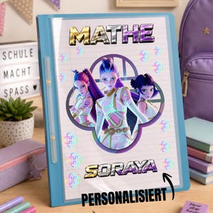 Könnte beinhalten: Ein hellblauer Schulordner mit dem Text MATHE und SORAYA in metallischer Schrift. Das Cover zeigt eine Grafik von drei animierten Figuren in weiß-violetten Outfits in einem kreisförmigen Rahmen, umgeben von kleinen holografischen Symbolen.