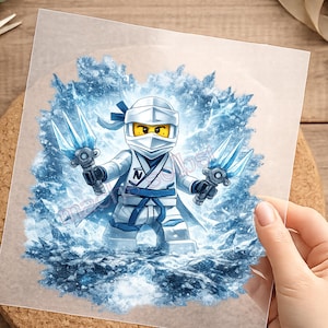 Könnte beinhalten: Ein weiß-blaues Grafik-Transferbild mit einer stilisierten Ninja-Figur, die eine Maske und eine Uniform mit einem Buchstaben N auf der Brust trägt. Die Figur hält zwei durchscheinende blaue, eisartige Waffen vor einem Hintergrund aus Eiskristallen.