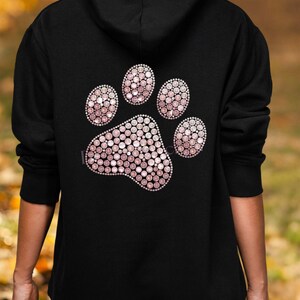Könnte beinhalten: Schwarzer Kapuzenpullover mit einem großen Pfotenabdruck-Design auf dem Rücken. Der Pfotenabdruck besteht aus kleinen, rosa und weißen Strasssteinen. Der Hoodie hat eine Kapuze und lange Ärmel. Der Text "Bügelsternchen" ist oben rechts sichtbar.