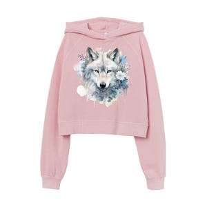 Könnte beinhalten: Hellrosa, kurz geschnittener Kapuzenpullover mit einem Aquarelldesign eines Wolfs, umgeben von blauen und weißen Blumen. Der Wolf hat graues Fell und blaue Augen. Der Hoodie hat lange Ärmel und eine Kapuze.