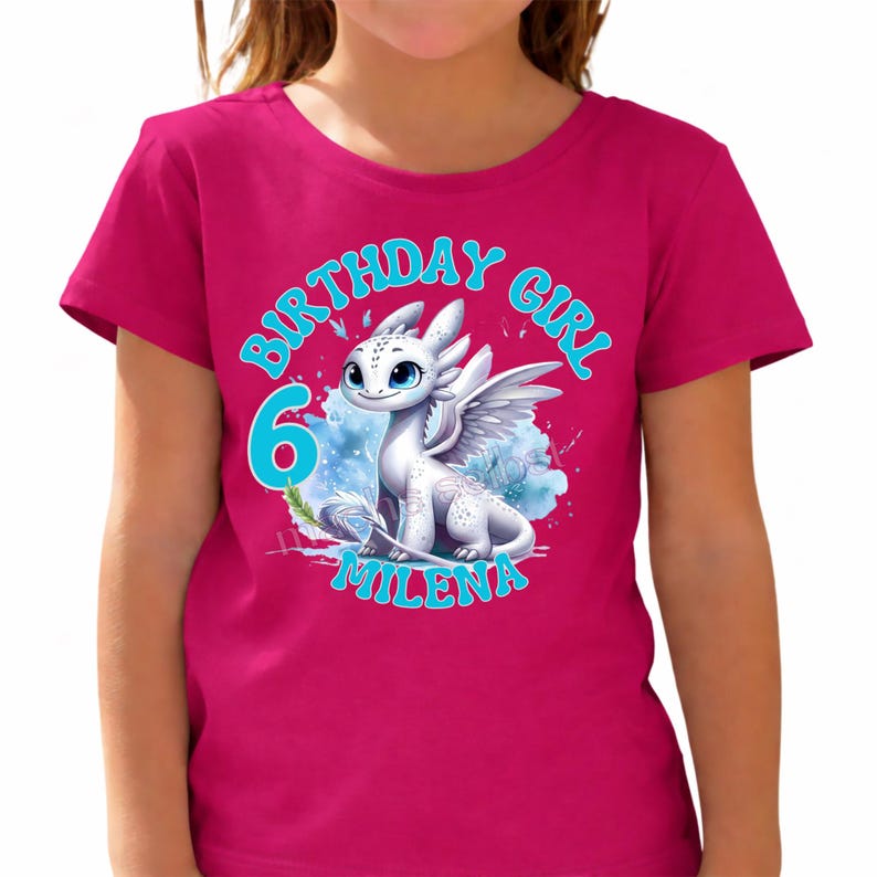 May include: A magenta t-shirt featuring a white dragon with blue eyes and wings. The text "BIRTHDAY GIRL 6 MILENA" is printed in blue, creating a festive design. The shirt is a children's apparel item.