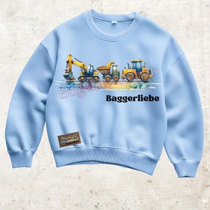 Könnte beinhalten: Hellblaues Sweatshirt mit einer Aquarellillustration von Baumaschinen: einem Bagger, einem Muldenkipper und einem Traktor. Der Text "Baggerliebe" ist darunter gedruckt. Ein kleines Patch mit Text befindet sich unten links.