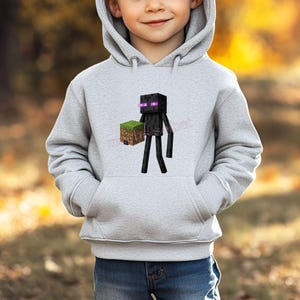 Könnte beinhalten: Hellgrauer Kapuzenpullover mit einem Minecraft Enderman-Motiv, der einen Block aus Erde und Gras hält. Der Enderman ist schwarz mit lila Augen. Der Pullover hat eine Vordertasche und eine Kapuze.