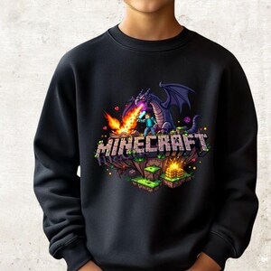 Könnte beinhalten: Ein schwarzes Langarm-Sweatshirt mit einem Grafikdruck einer Minecraft-Szene. Das Design zeigt einen violetten Drachen, der Feuer auf eine Figur mit Schwert speit, sowie das Wort MINECRAFT in Blockbuchstaben über pixeligen Geländeblöcken.