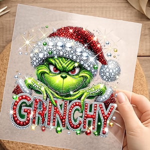 Könnte beinhalten: Ein Diamant-Malerei-Aufkleber mit einer grünen Kreatur mit roten Augen, die eine rot-weiße Weihnachtsmütze trägt, akzentuiert mit funkelnden Strasssteinen. Das Wort GRINCHY steht in großen, roten, mit Strass besetzten Buchstaben.