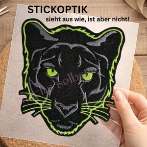 Könnte beinhalten: Ein gestickter Aufnäher mit einem schwarzen Pantherkopf, leuchtend hellgrünen Augen und einer hellgrünen Umrandung. Der Aufnäher enthält den Text STICKOPTIK und sieht aus wie, ist aber nicht! in schwarzer Schrift auf der transparenten Folie.