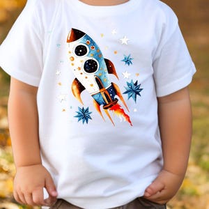 Könnte beinhalten: Weißes T-Shirt mit einem bunten Raketendesign in Blau, Orange und Weiß, umgeben von Sternen. Die Rakete hat zwei große runde Fenster. Das T-Shirt ist für Kinder.