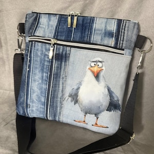 Könnte beinhalten: Jeans-Umhängetasche mit einer Cartoon-Möwen-Illustration. Die Tasche hat einen schwarzen, verstellbaren Riemen, silberne Reißverschlüsse und ein hellblaues Jeans-Design. Die Möwe ist grau, weiß und orange.