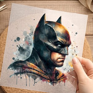 Könnte beinhalten: Ein Aquarelldruck von Batman im Profil, mit dunkler Kapuze und ernstem Gesichtsausdruck. Das Kunstwerk verwendet Schwarz-, Blau- und Gelbtöne mit Farbspritzern im Hintergrund. Der Druck wird von einer Hand gehalten.