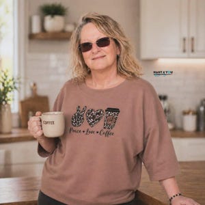 Könnte beinhalten: Ein hellbraunes Kurzarmshirt mit einer Grafik aus einem Leopardenmuster-Friedenszeichen, einem Herz und einem Kaffeebecher. Unter der Grafik steht der Text Peace Love Coffee. Das Shirt wird mit einer beigen Tasse mit der Aufschrift COFFEE kombiniert.